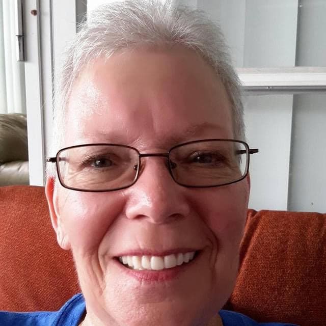 Smiling woman with short grey hair and glasses wearing a blue shirt against orange upholstery.